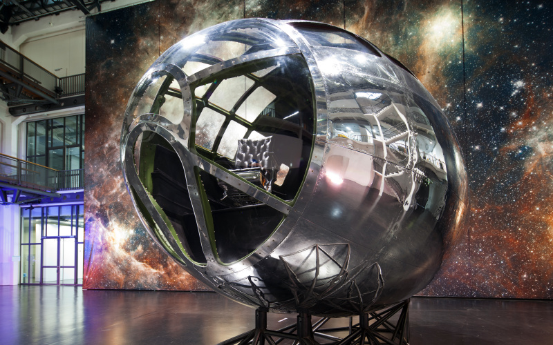 Zu sehen ist ein silbernes Gestellt, das an eine Raketenspitze erinnert und vor einer großen Leinwand mit dem Motiv einer Galaxie steht. In der vorderen Glaskuppel des Gestellt ist ein schwarzer Ledersessel zu erkennen. 