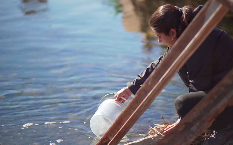 Einzelbild eines der beiden Videos des Werkes Hidroscopia Loa. Man sieht wie die Künstlerin Claudia González Godoy eine Wasserprobe aus dem Fluss Loa entnimmt.
