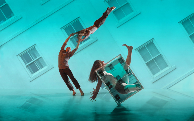 You can see 3 dancers in front of a turquoise-colored, slanted house facade.