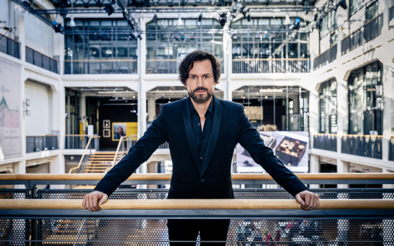 The picture shows Ole Scheeren. He is standing in a black suit at the ZKM, on one of the bridges above the foyer against the background of the museum balcony. He forms the center of the image and looks directly into the camera. 