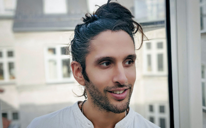 In the picture you can see the designer and artist Cyrus Clarke. He wears his black long hair in a bun and looks slightly down to the right.