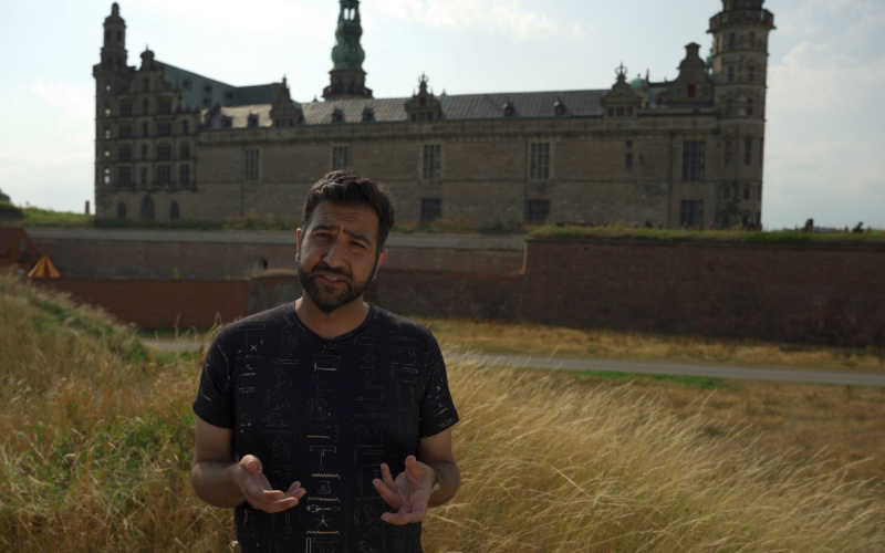 Wassim Z. Alsini is shown in the picture. He is standing outside in a green landscape and has a castle in the background. He is looking directly into the camera and was portrayed while speaking.