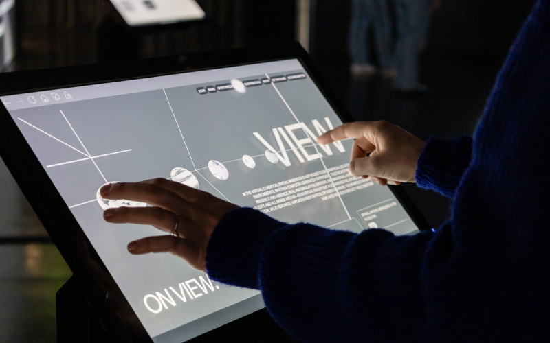 Foto eines Displays in der Ausstellung »Matter. Non-Matter. Anti-Matter«. Auf dem Display steht "View" und es sind kleine planetenartige Symbole zu sehen. 