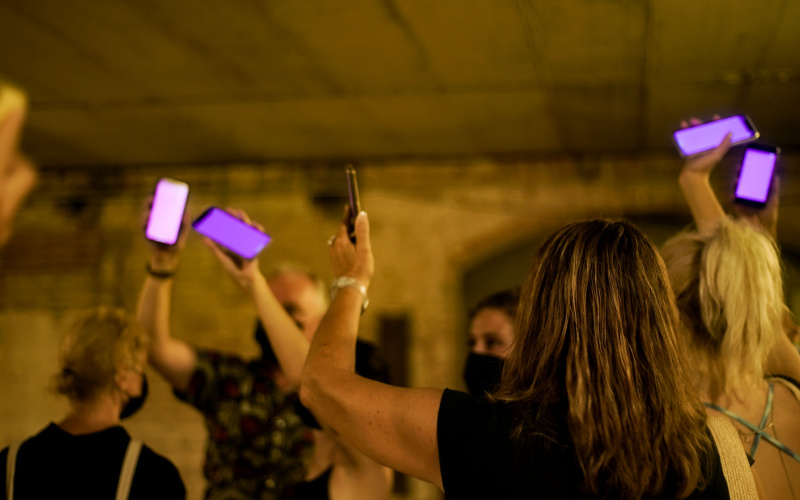 Zu sehen sind mehrere Personen in einem abgedunkelten Raum, die ihre Arme nach oben strecken. In ihrer Hand halten sie ein Smartphone, dass pink aufleuchtet.