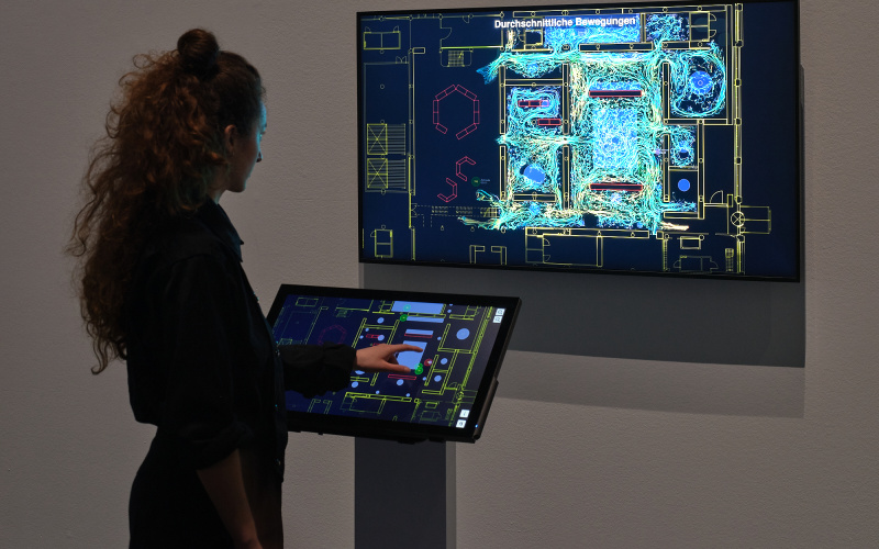 »TRACES« von Bernd Lintermann. Zu sehen ist eine Person vor einem Bildschirm, auf dem verschiedenfarbige Linien abgebildet werden. Vor der Person befindet sich ebenfalls ein Touchscreen-Bildschirm, auf dem sich ebenfalls Linien befinden. 