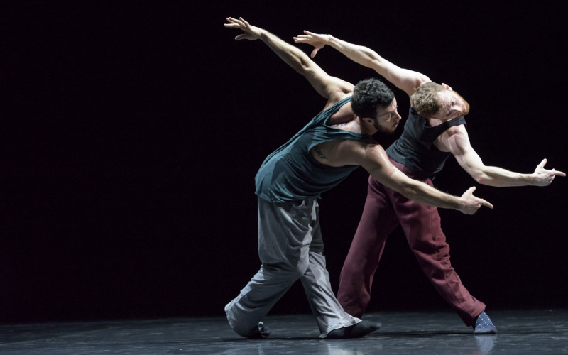 Zwei Tänzer der Choreografie »Friends of Forsythe«. Sie stehen zusammen auf einer schwarzen Bühne und verrenken ihre Körper kunstvoll parallel zueinander.