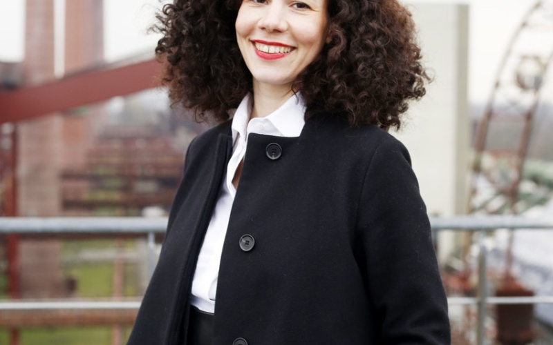 Jasmin Grimm in a black coat and white blouse standing outside in factory area