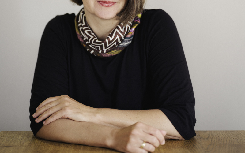 Portrait of Elke Buhr sitting at a wooden table in front of a white wall