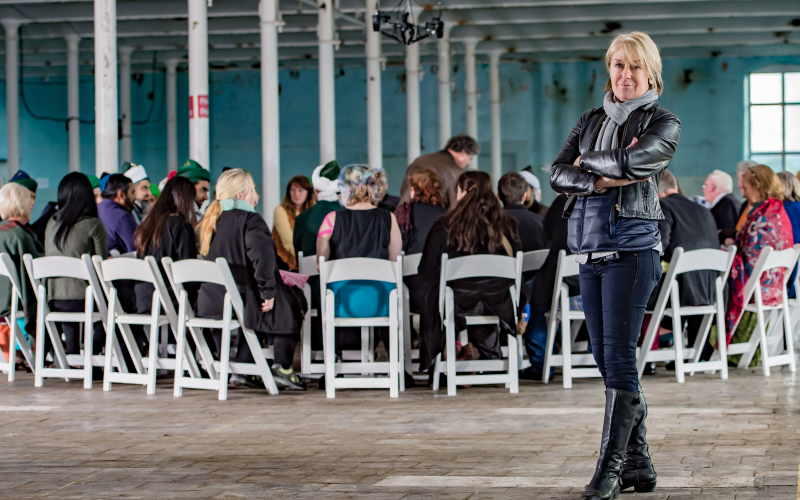 In the foreground is the artist Suzanne Lacy and in the background you can see many people sitting together in a circle of chairs.
