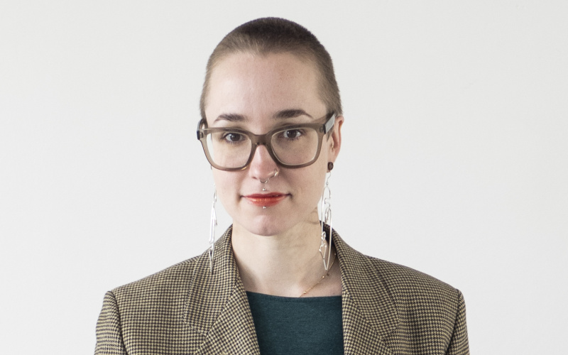 A young woman with glasses stands in front of a white background and wears a beige plaid coat.