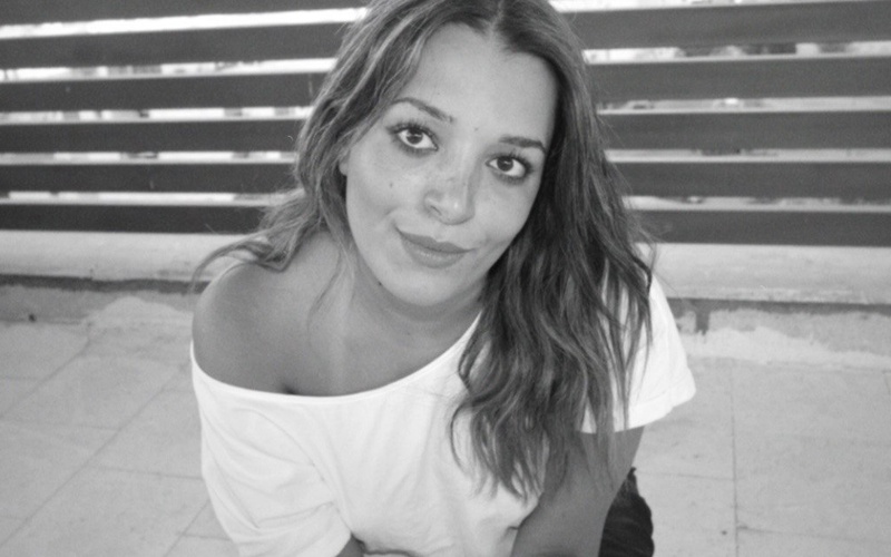 Lächelnde Person mit weißem Shirt (Jana Hammoudeh) sitzt vor einem Zaun.