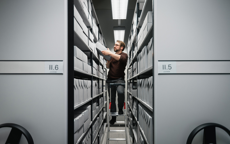 One person stands on a ladder and puts away archive boxes.