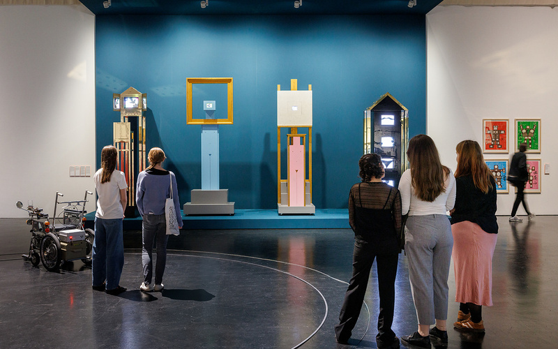 Blick in die Ausstellung The Story That Never Ends, 5 Personen schauen Kunstwerke an, blauer Hintergrund
