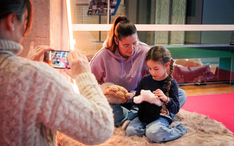 Eine Person macht ein Foto von einer Mutter mit Kind, die mit einem Roboter-Meerschweinchen schmusen.