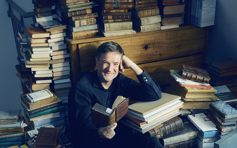 Der Autor Markus Orths sitzt lächelnd zwischen Bücherstapeln