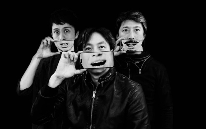 Three people in a white-and-black photo are each holding a cell phone in front of the lower half of their faces.