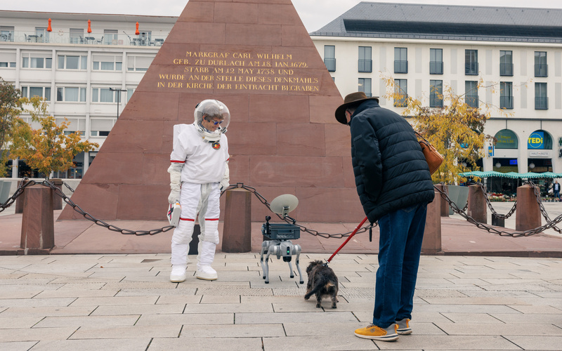 Das Bild zeigt eine Person in einem Raumanzug, die zusammen mit einem Roboterhund einen Hund und dessen Begleiter begrüßt.