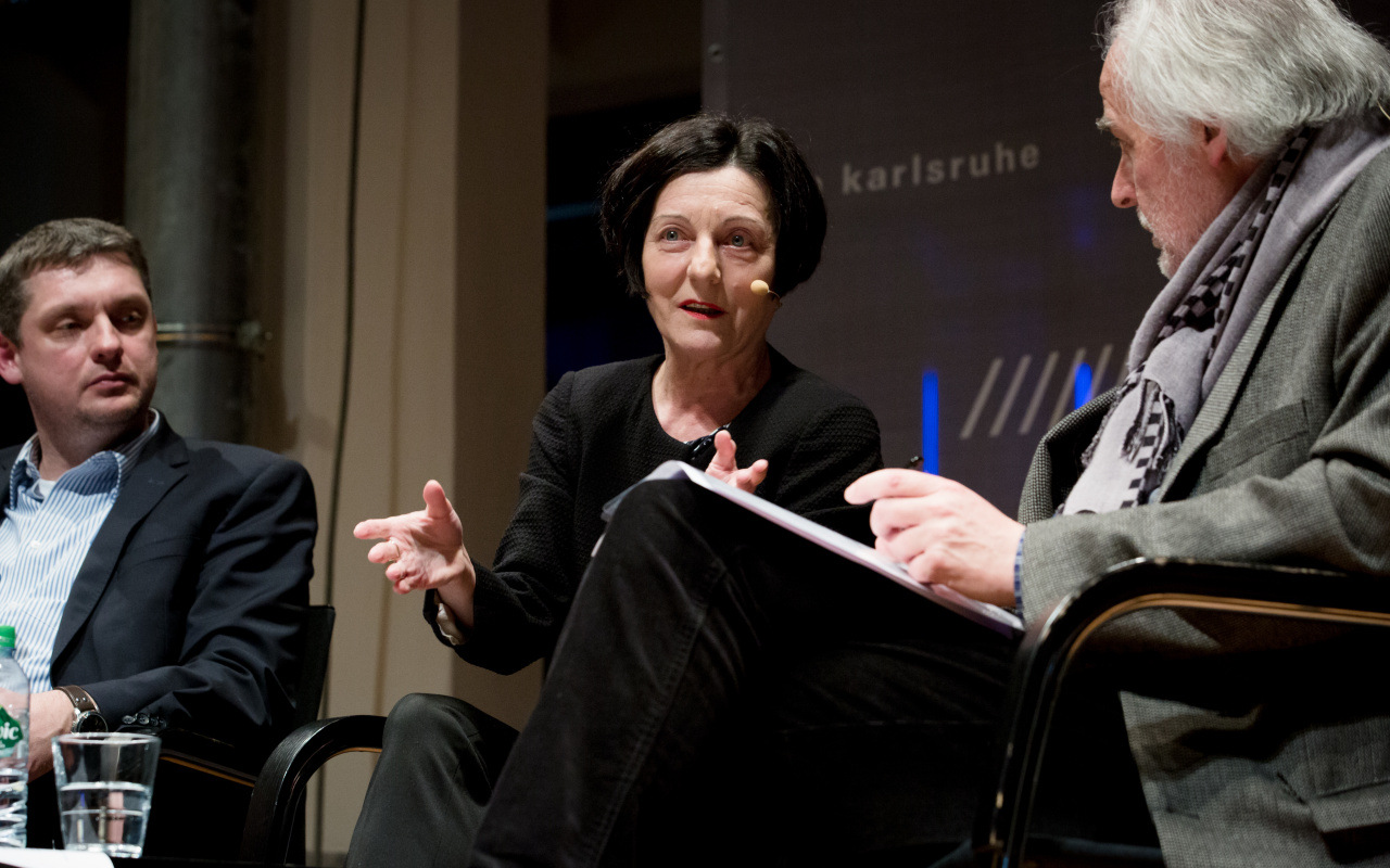 Three people are sitting on a stage while the one in the middle, a woman, is talking