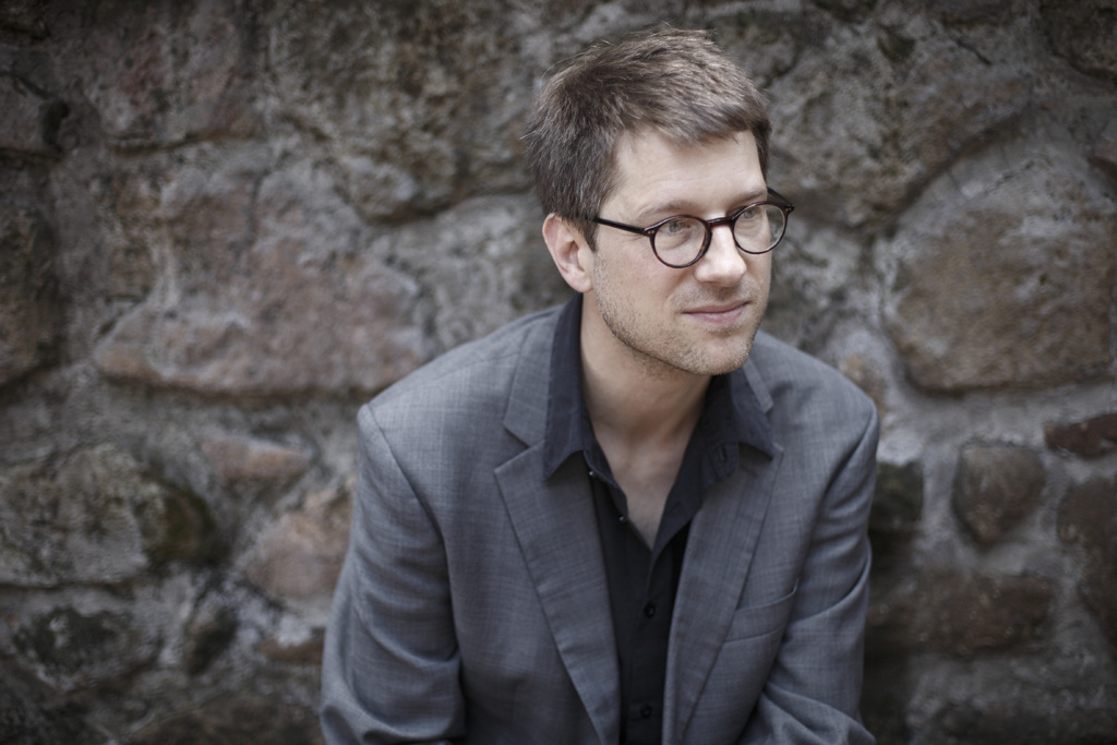A man with glasses sits in front of a stone wall