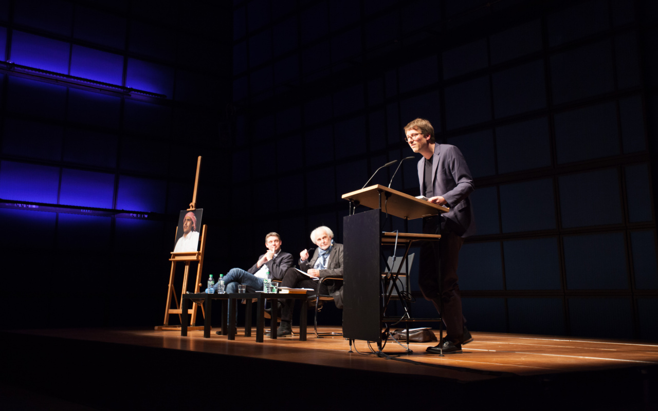 Two men are sitting on a stage, a third is standing a desk and talks