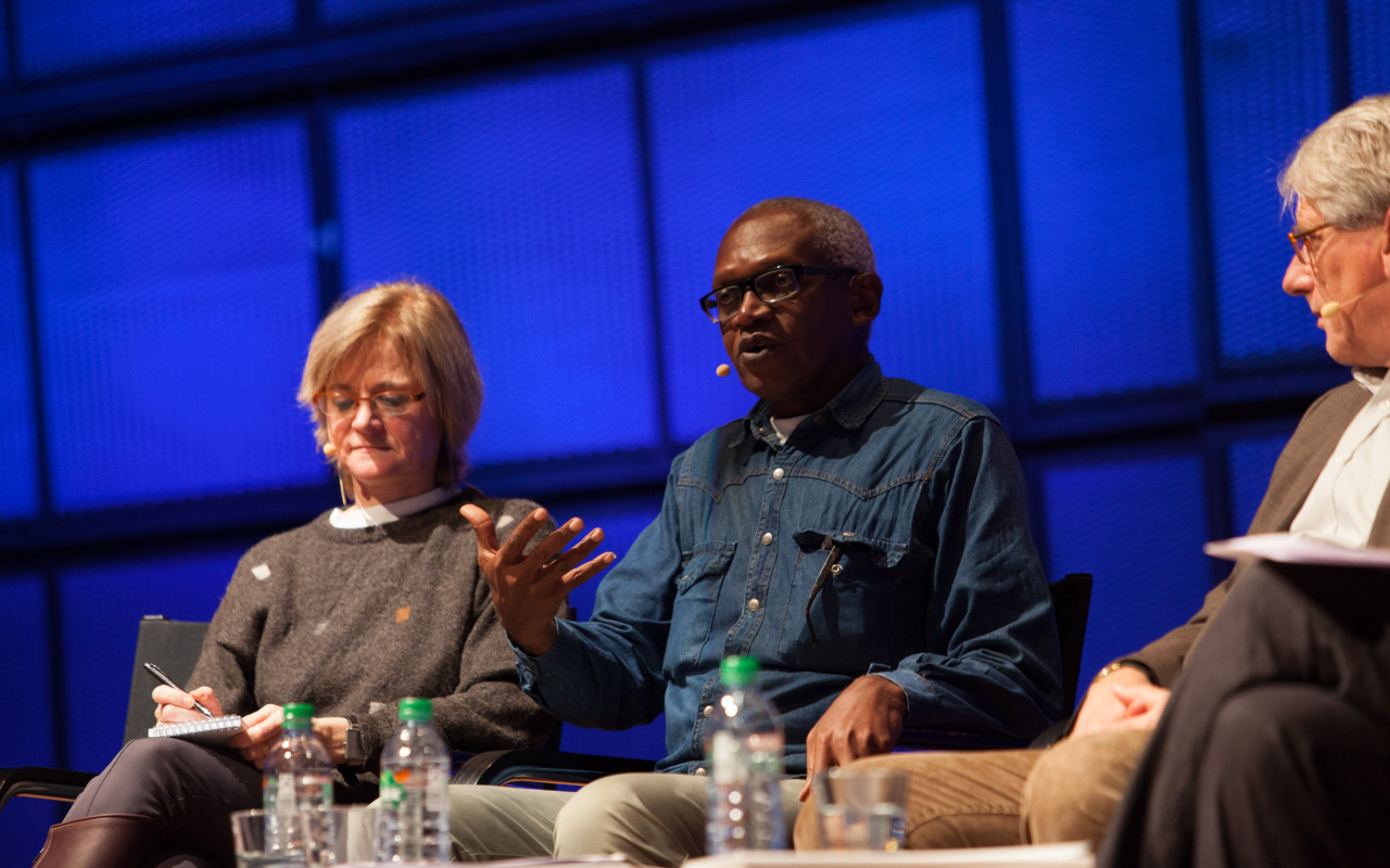 Three people are sitting on a stage while one of them, a man, is talking