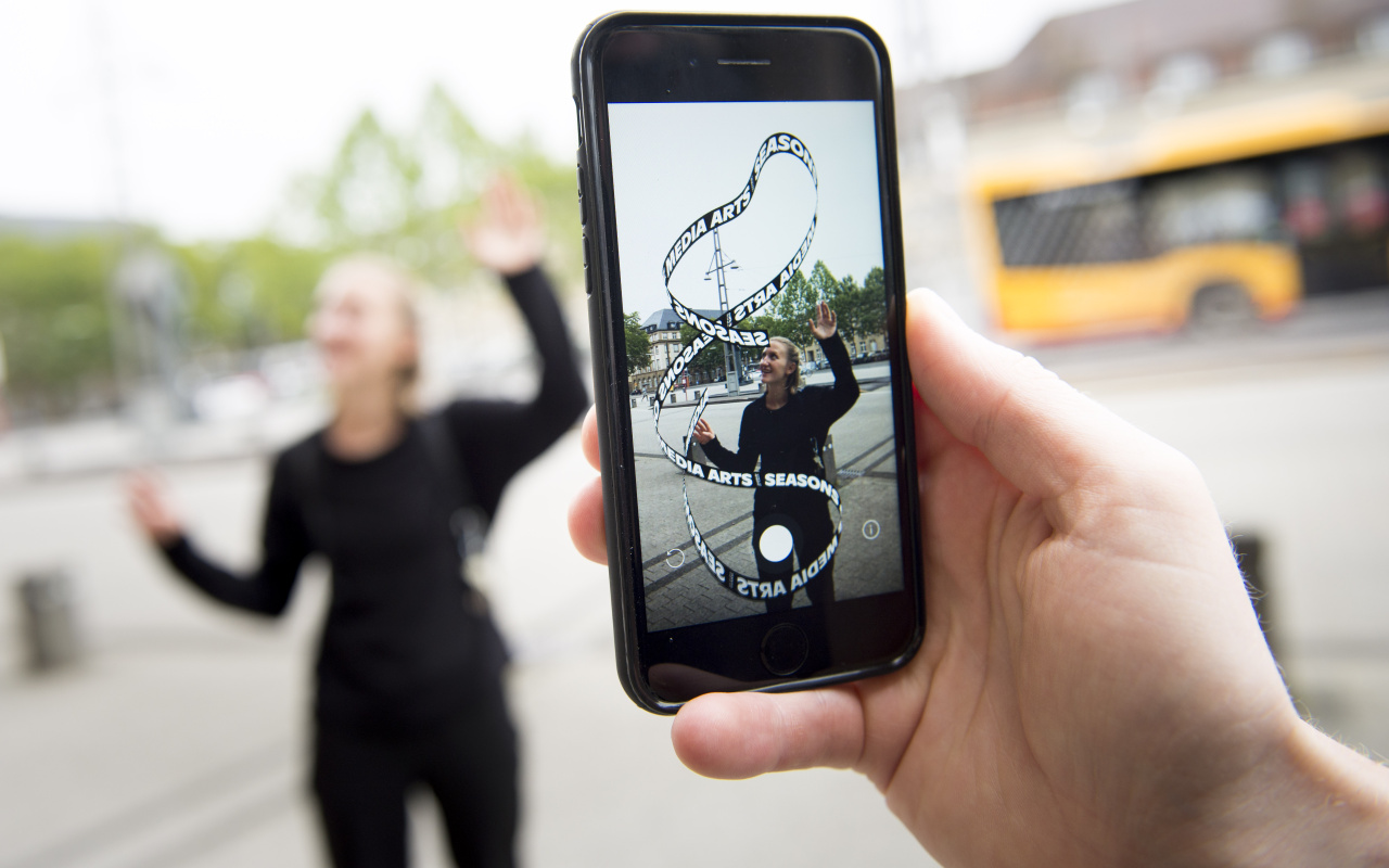 Im Vordergrund sieht man ein Handy auf dessen Display ein App-Kunstwerk zu sehen ist. Die virtuelle »Seasons of Media Arts« Schlaufe und eine blonde Frau, die die Enden der Schlaufe versucht zusammenzuhalten.