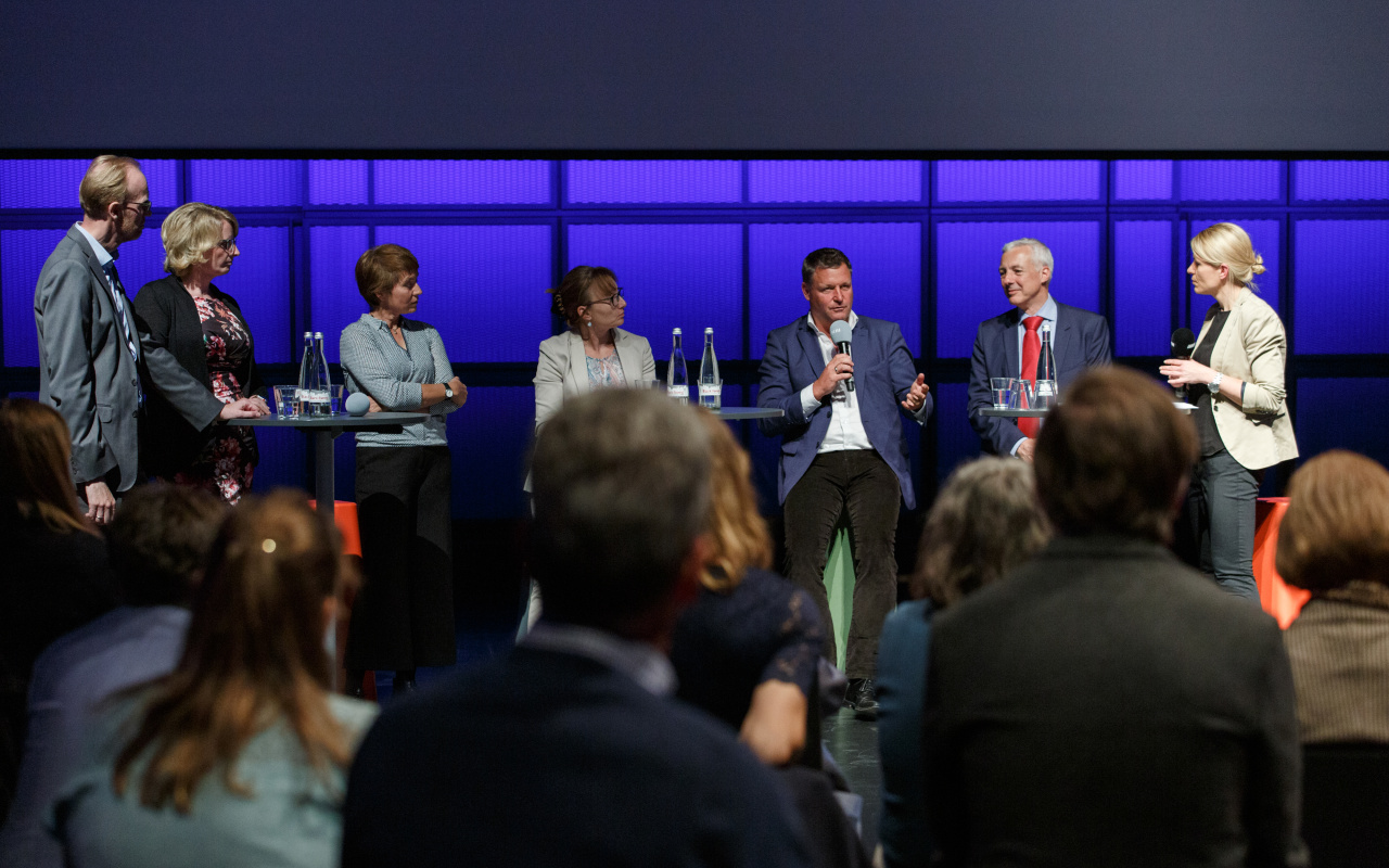 Vor einer Gruppe Menschen diskutieren sieben Personen an Stehtischen mit Mikrofonen in der Hand.