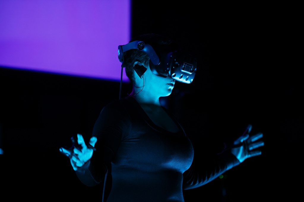 Person mit VR Brille und 2 Controllern auf einer Blau beleuchteten Bühne.