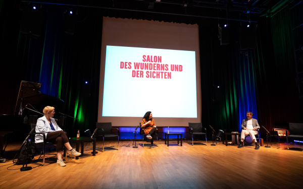 The dramaturges of the Munich Biennale sit in a semicircle at a distance. Behind them is a projection on which »Salon of wonder and views« is written.