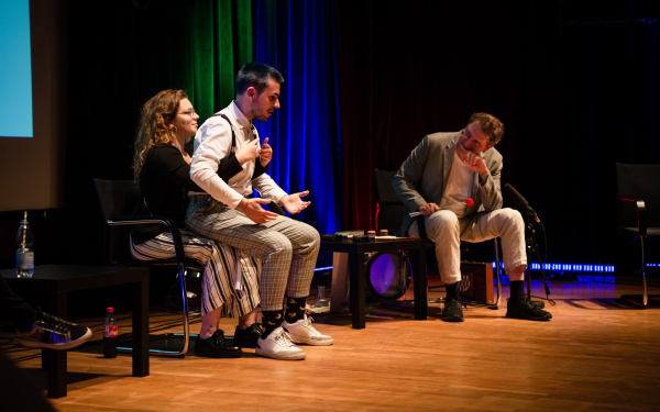 Óscar Escudero sits on the lap of Belenish Moreno-Gil and speaks. Belenish gestures with her hands so that they resemble a hand puppet piece. Manos Tsangaris listens to him by leaning over towards the duo.