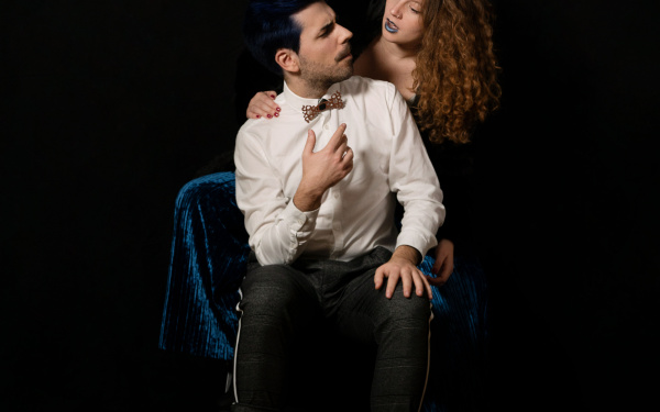 A young man is sitting on a chair. His head is turned to the side and he looks at a young woman sitting behind him. Her hand is resting on his shoulder.