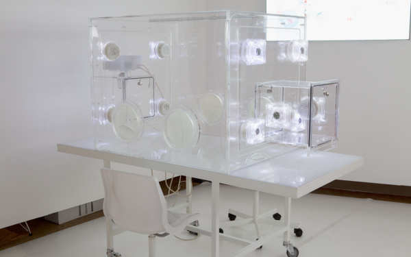 Leerer Baby Brutkasten auf einem rollbaren weißen Tisch. Zwei weiße Stühle stehen an dem Tisch, im Hintergrund läuft eine Projektion auf der weißen Wand.
