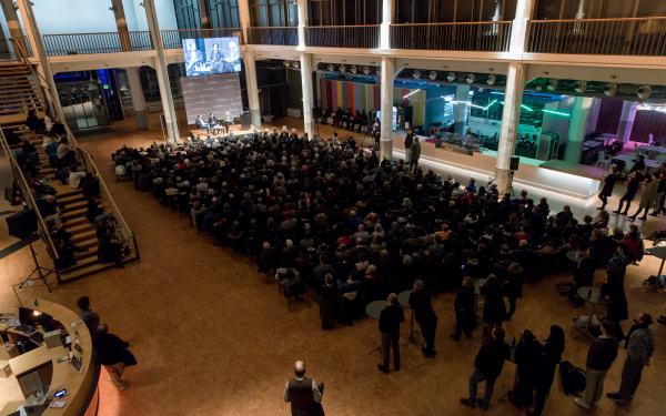 Menschen sitzen vor eine Bühne, über der eine große Leinwand hängt