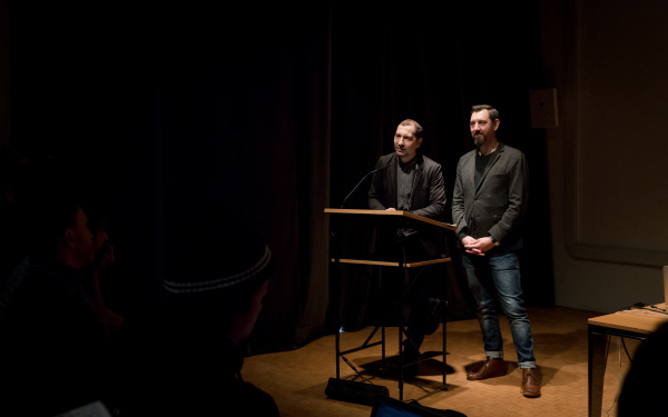 Zwo persons stand at a desk and talk to the audience