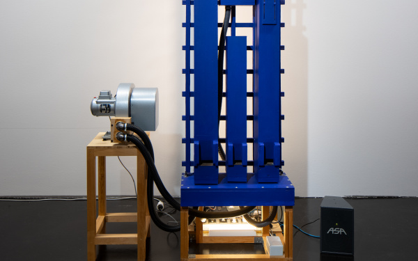Zu sehen ist eine rechteckige, maschinenartige Installation, welche auf einem Hocker steht. Links neben der Installation steht ebenfalls ein kleiner Hocker, auf welchem ein Objekt liegt. 