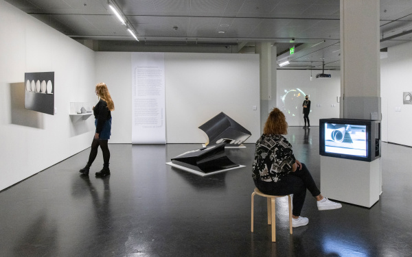 Drei Frauen in der Ausstellung. Die Frau links betrachtet eine Skulptur, die an die Wand gehängt ist, die Frau rechts schaut einen Film in einem Fernseher, in der Mitte sind abstrakte Figuren, an der hinteren Wand hängt eine Tapetenrolle mit Text darauf.