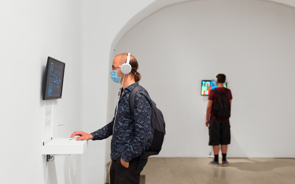 Ein Mann steht vor einem Bildschirm, der an der Wand hängt. Er nutzt die Computermaus, die unter dem Bildschirm auf einem Tisch liegt.
