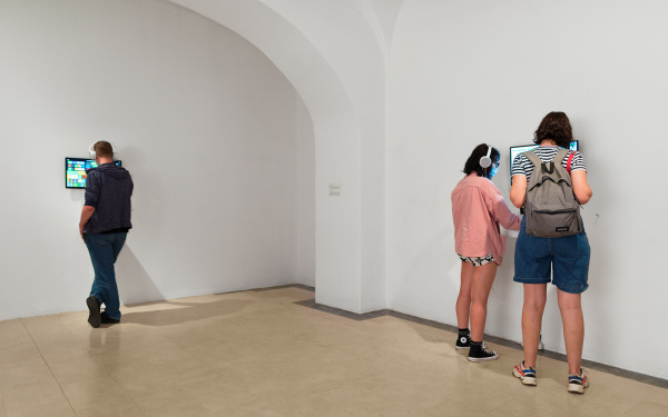 Menschen stehen an Monitoren, die an der Wand befestigt sind und tragen dabei Kopfhörer.