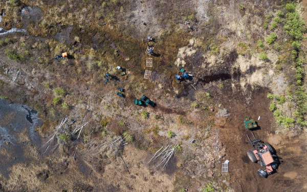 A drones shot from above, looking at a swamp landscape. People work on it with garbage bags, excavators and pallets.