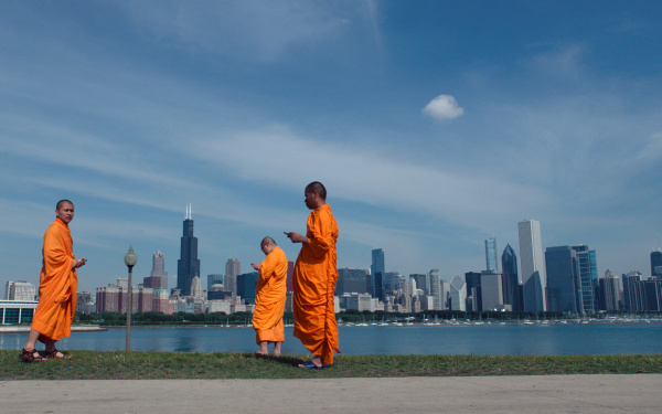 Drei Männer in tibetischer Tracht stehen vor der Uferskyline einer großen modernen Stadt. Alle drei schauen auf ihr Smartphone. 