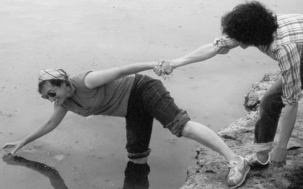 Lynn Margulis steht mit einem Bein im Wasser und hält den Finger einer Hand ins Wasser. Mit dem anderen Bein steht sie auf der Erde und die andere Hand hält eine fremde Hand fest. Sie steht gekippt und lacht.