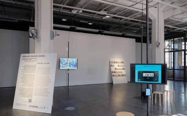 Blick in die Ausstellung. Zu sehen sind eine große Schrifttafel und zwei im Raum stehende Bildschirme. Vier Stühle stehen im Raum verteilt.