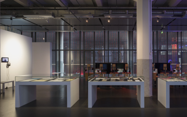 Ein weiter Blick in die Ausstellung. Vorne Vitirinentische, links eine Stellwand mit Bildschirmen und Kopfhörern, im Hintergrund mehr Bildschirme und Stühle.