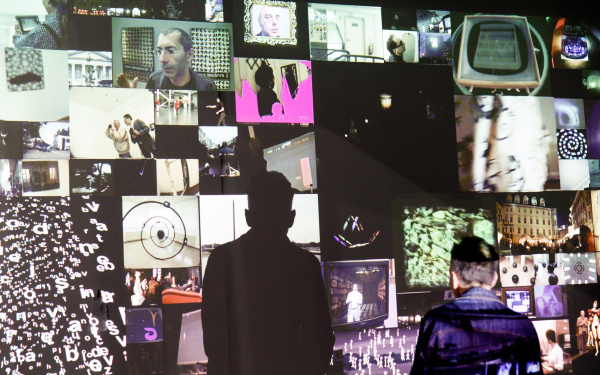 Zu sehen ist eine Video-Installation. Es werden viele kleinere Bilder auf eine Wand projiziert, wobei ein Mosaikartiges Bild entsteht. Davor steht ein betrachtender Mann. Er wirft einen auffälligen Schatten auf die Wand.