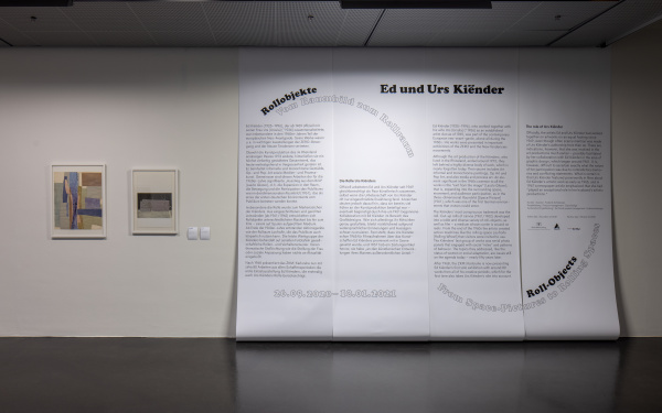 Neben großflächig montiertem Ausstellungstext an der Wand sind auf dem Bild noch zwei weitere Werke an der wand aufgehängt und gerahmt zu sehen.