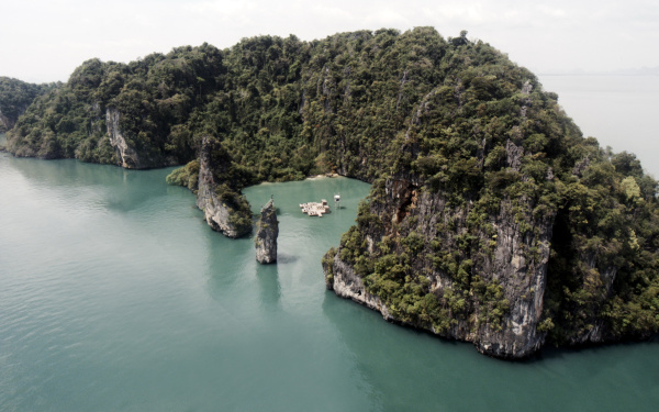 Das Bild zeigt eine von Wasser umgebene kleine Insel. In der Mitte des Bildes ist eine kleine Bucht. In dieser Bucht liegt ein Plateau mit zwei kleinen Bauten darauf, das auch rundherum von Wasser umgeben ist.