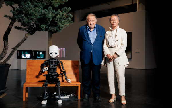 Zu sehen ist ein Roboter, dessen Aufbau dem Korpus des Menschen gleicht. Er sitzt auf einer Bank. Recht daneben steht Peter Weibel zusammen mit Soun-Gui Kim.