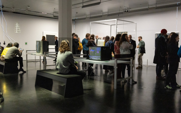Ausstellungsansicht »Matter. Non-Matter. Anti-Matter« im ZKM | Zentrum für Kunst und Medien Karlsruhe, 2022. Eine Person trägt Kopfhörer und sitzt vor einem Röhrenfernseher. Im Hintergrund befindet sich eine Menschentraube.