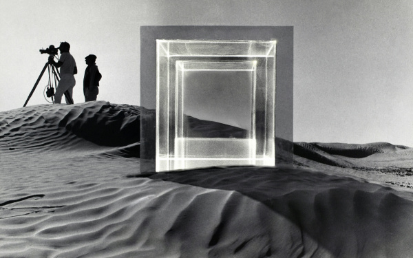 In a desert landscape around 1968, filming of the movie "Tele-Mack" took place. In the foreground is a large square glass sculpture and in the background are two men with a film camera. The picture is black and white.
