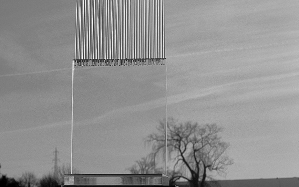 In the middle of a landscape stands a large glass sculpture. The lower part of the sculpture is made of transparent glass and the upper part is made of glass, which looks like a harp and has no clear edge upwards. The image is black and white.